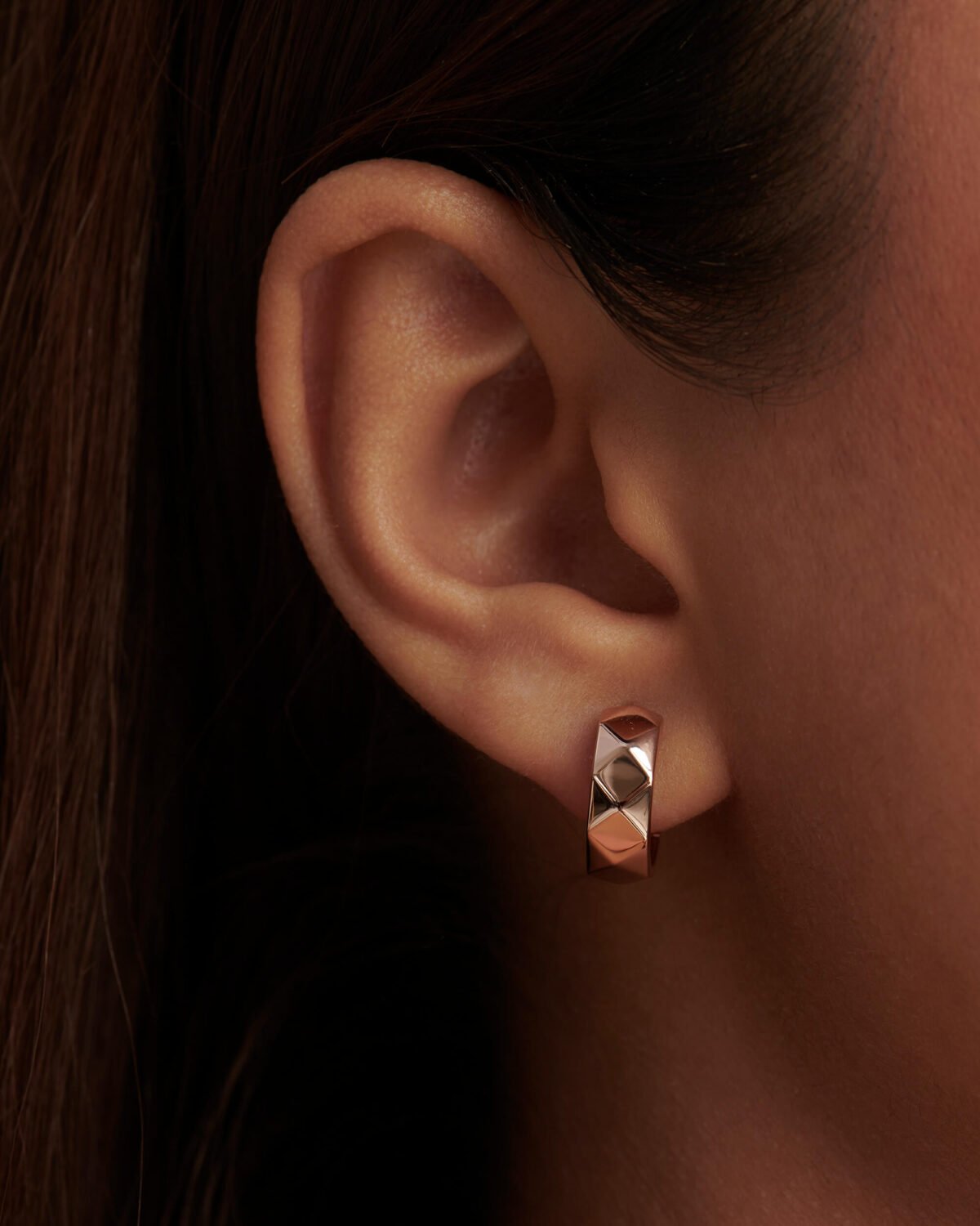 Close-up of an ear wearing a geometric rose gold earring with dark hair and skin.